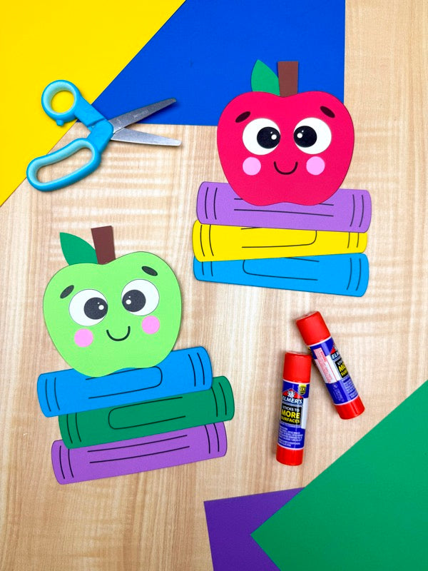 Colorful paper craft of apples and books with scissors and glue sticks on a wooden surface.