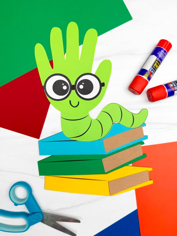 Handmade paper craft of a green caterpillar with googly eyes on books, surrounded by construction paper and craft supplies.