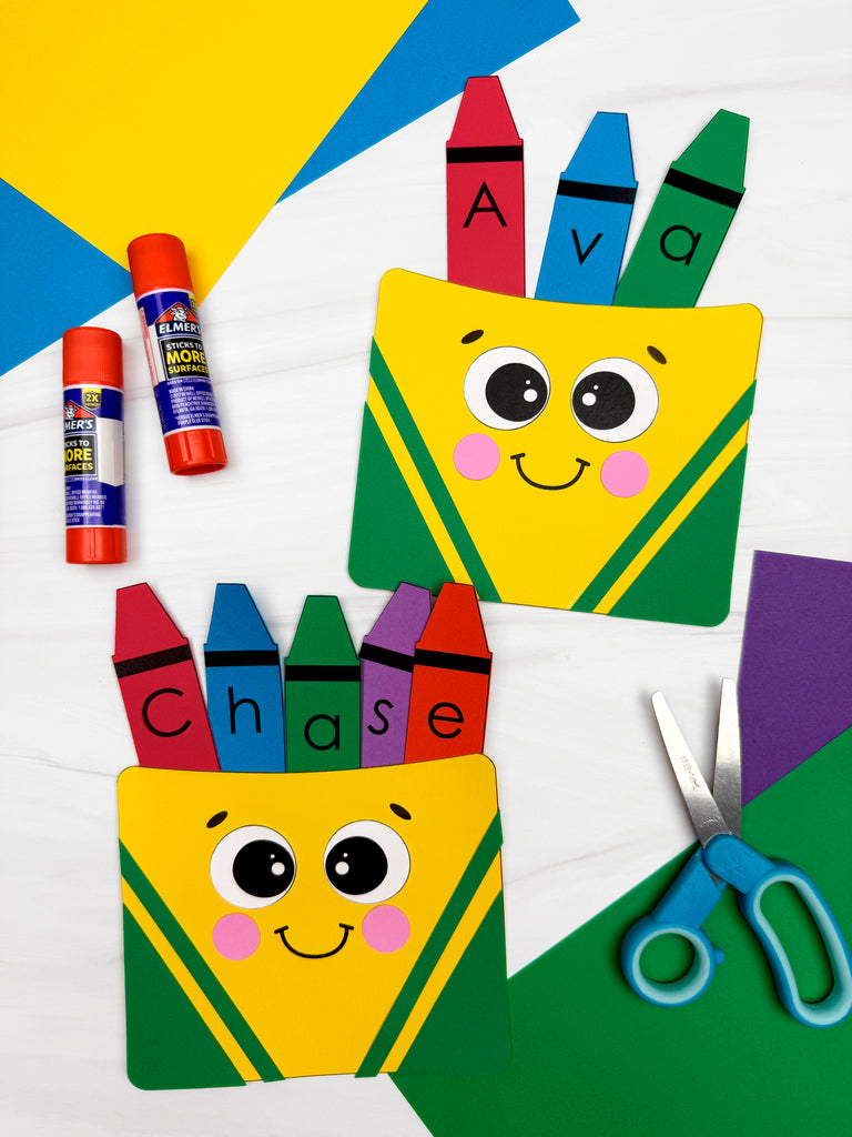 Two colorful paper crafts shaped like crayon boxes with smiling cartoon faces hold crayon cutouts labeled with the names "Ava" and "Chase" in large letters. Craft supplies including glue sticks, scissors, and colored paper are scattered around the scene on a white surface.