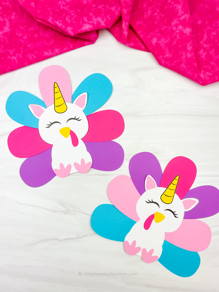 Two colorful unicorn turkey disguise crafts on a white surface with a pink fabric background.