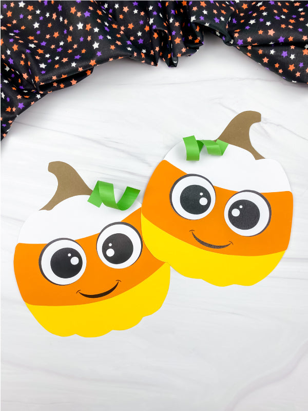 Two smiling candy corn pumpkin paper crafts with large eyes, brown stems, and green paper curls are displayed on a white surface beneath Halloween star-patterned fabric.