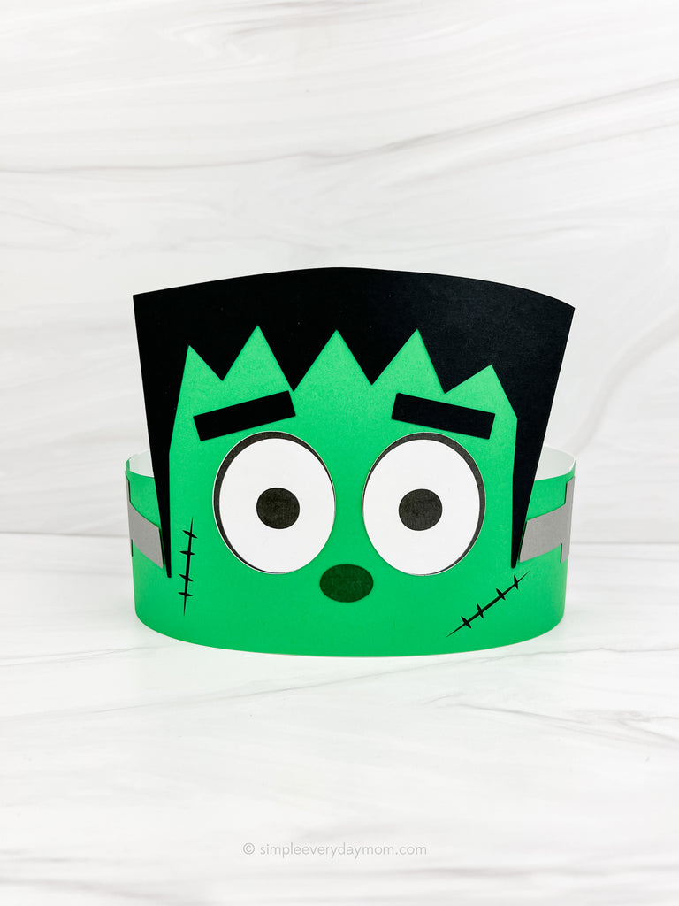 A green and black Frankenstein-themed paper party hat with large cartoon eyes, shown on a white marble-textured background.
