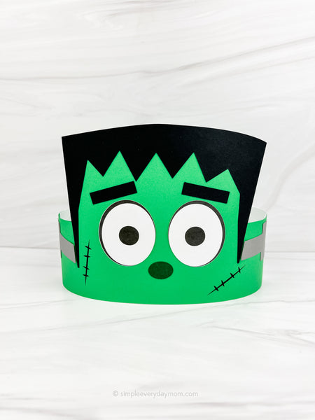 A green and black Frankenstein-themed paper party hat with large cartoon eyes, shown on a white marble-textured background.