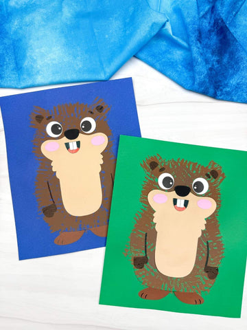 Two fork painted groundhog crafts, one on blue paper and one on green paper, with a blue fabric background.