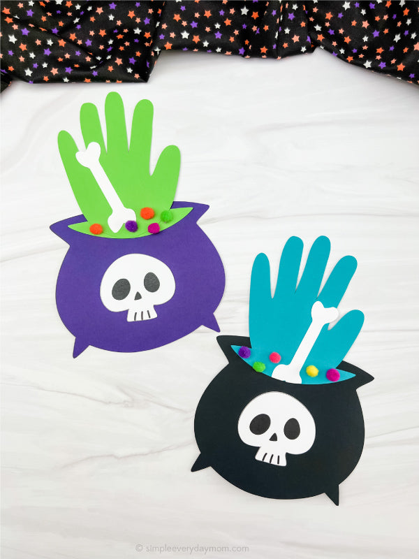Two Halloween craft decorations shaped like cauldrons with skull faces, one purple and one black, each holding a hand-shaped green and blue element with a white bone and small multicolored pom-poms, displayed on a white fabric background.