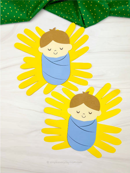 Two Christmas handprint manger crafts featuring baby Jesus with brown hair, closed eyes, and a blue swaddling blanket surrounded by yellow paper handprints representing hay. Both crafts are displayed on a white wooden surface with green star-patterned fabric.