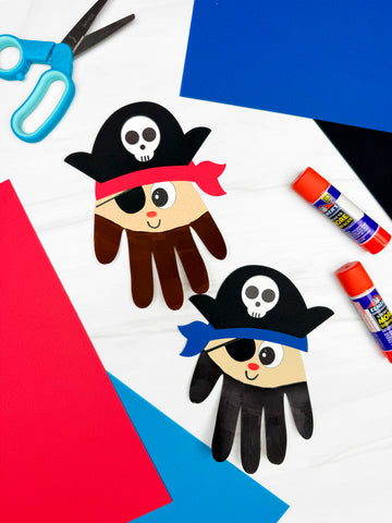 2 handprint pirate crafts with glue sticks and scissors in the background. Colored paper along the corners.