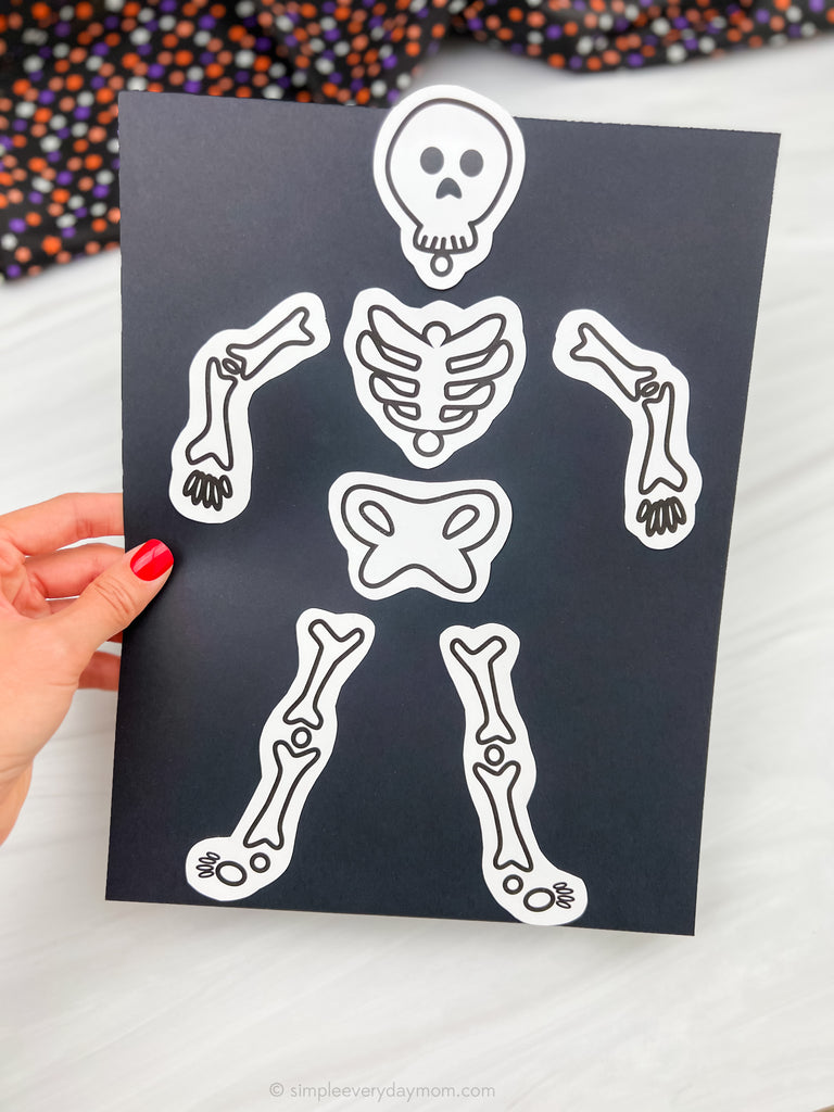 A hand with red nail polish holds a black sheet featuring white cut-out skeleton bone pieces, shown against a light marble background with a colorful patterned fabric blurred in the top corner.