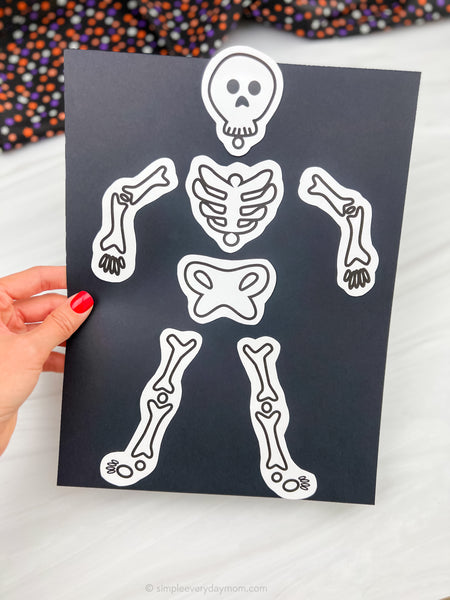 A hand with red nail polish holds a black sheet featuring white cut-out skeleton bone pieces, shown against a light marble background with a colorful patterned fabric blurred in the top corner.