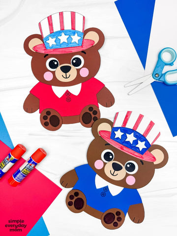 Two paper bear crafts with American flag hats on a white background with scissors and glue sticks.