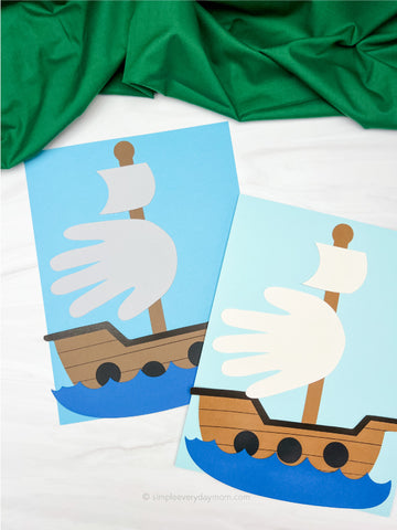 Two handprint crafts shaped like ships on a white background with green fabric.