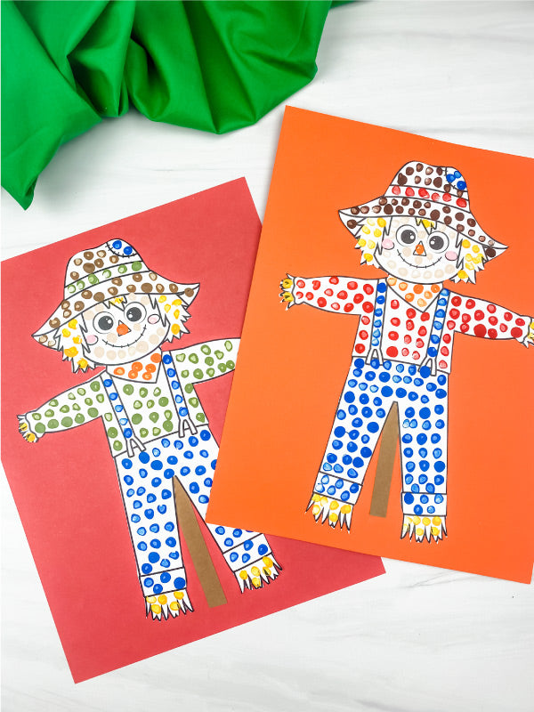 Two colorful paper scarecrow crafts on red and orange papers, with a green fabric roll in the corner, photographed on a white surface.