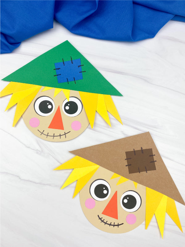 Two paper craft scarecrow faces on a white background with blue fabric.