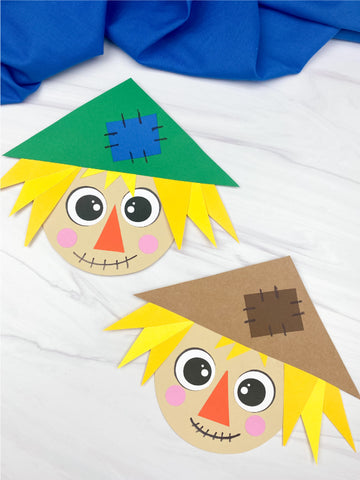 Two paper craft scarecrow faces on a white background with blue fabric.