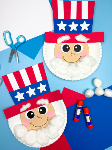 Two paper plate crafts resembling Uncle Sam's face, each decorated with a red, white, and blue top hat, cotton balls for hair and beard, and large cartoon eyes and a smile. Art supplies like scissors, glue sticks, and cotton balls are scattered around on a blue and white background.