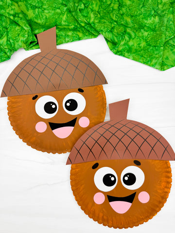 Two brown paper plate acorns with googly eyes and pink cheeks on a marble background with green fabric