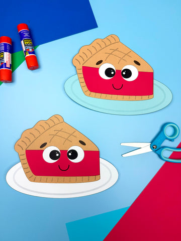 Two paper pie crafts with faces on a blue background with glue and scissors.