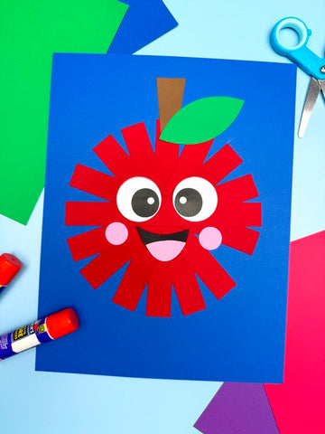 Child's craft project of a paper strip red apple with big eyes on blue paper, surrounded by colorful cutouts and craft supplies.