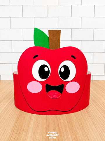 A red apple-shaped paper headband craft with a cute cartoon face, pink cheeks, and a green leaf on a wooden table against a white subway tile wall. A small 'simply everyday mom' watermark is visible at the bottom.