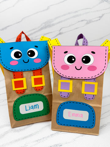 Two colorful backpack paper bags with cartoon faces and name labels on a marble surface