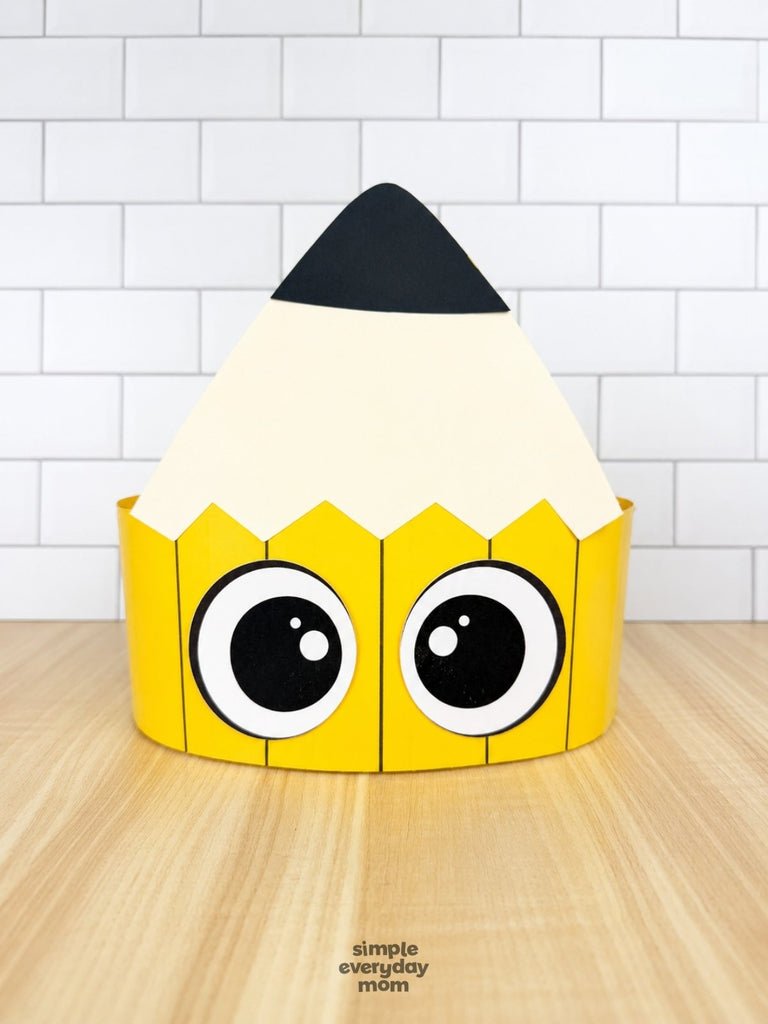 A cute, cartoon-style pencil-shaped paper crown with large expressive eyes on the yellow brim and a black eraser tip, displayed on a light wood surface against a white subway tile background.