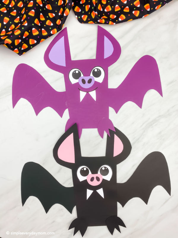 Two cute paper bat crafts stacked on top of each other, with one purple bat above a black bat. The purple bat has pink inner ears and a pink nose, and the black bat has pink inner ears and a pink nose.