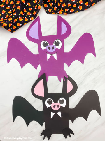 Two cute paper bat crafts stacked on top of each other, with one purple bat above a black bat. The purple bat has pink inner ears and a pink nose, and the black bat has pink inner ears and a pink nose.