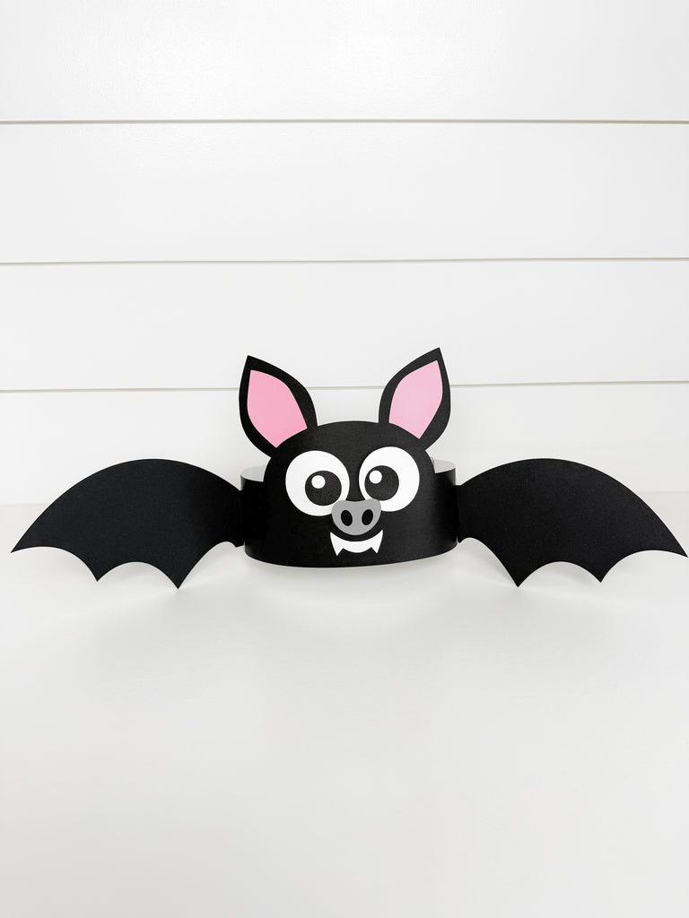 A black bat costume headband with pink inner ears, large cartoon eyes, and small fangs, shown against a white paneled background.