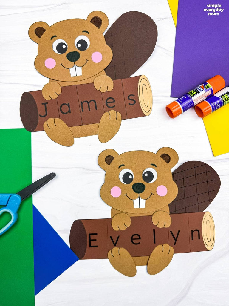 Two beaver name crafts on marble background with colored paper, glue sticks, and scissors 