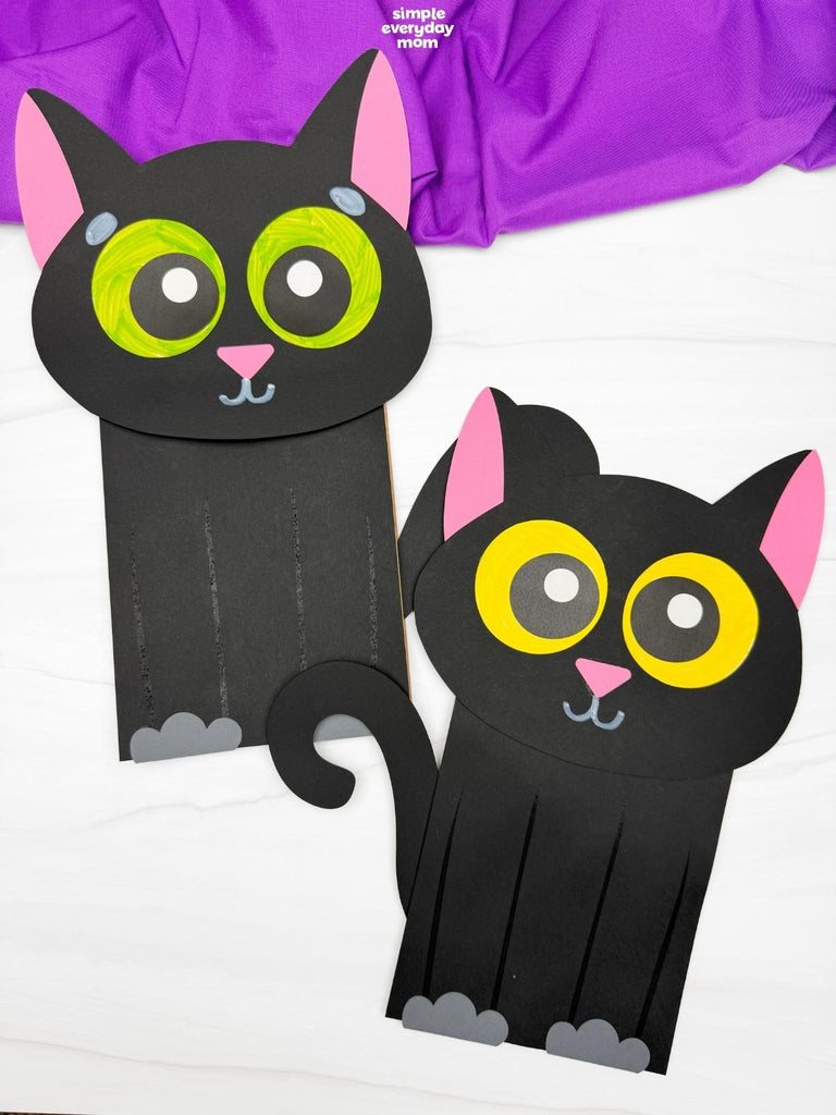 Two black cat paper bag puppet on white surface with purple fabric on top