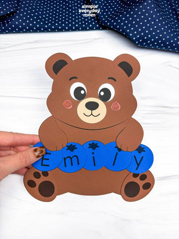 A hand holds a brown bear name craft. The background shows a white textured surface with a navy blue polka dot fabric at the top. 
