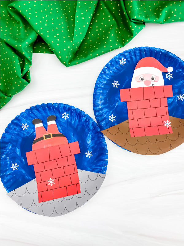 Two paper plate crafts shaped like Santa Claus in a chimney on a white background with green fabric.