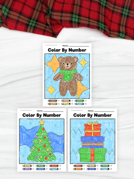 Color by number activity sheets featuring a bear, Christmas tree, and presents on a white surface with plaid fabric.