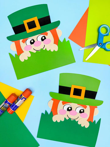 Two paper crafts of leprechauns with green hats on a blue background, surrounded by craft supplies.