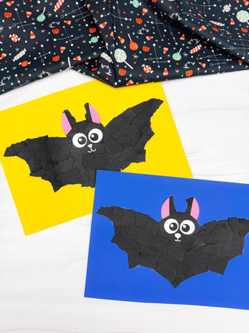 Two torn paper bat crafts on yellow and blue paper, with a folded black fabric featuring candy and moon pattern in the background, arranged on a white wood surface.