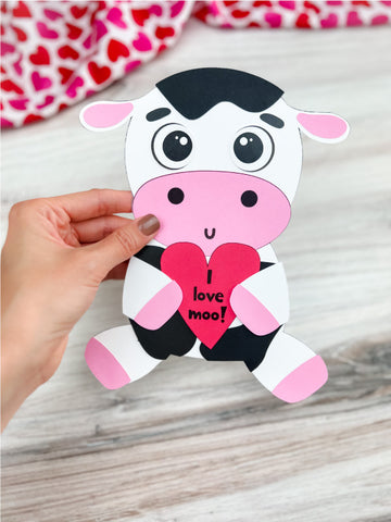 Hand holding a paper cow craft with a heart that says 'I love moo!' on a wooden surface.