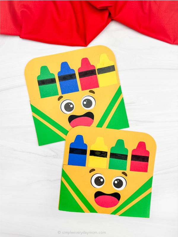 Two colorful cardstock crafts shaped like crayon boxes with faces on a white background.
