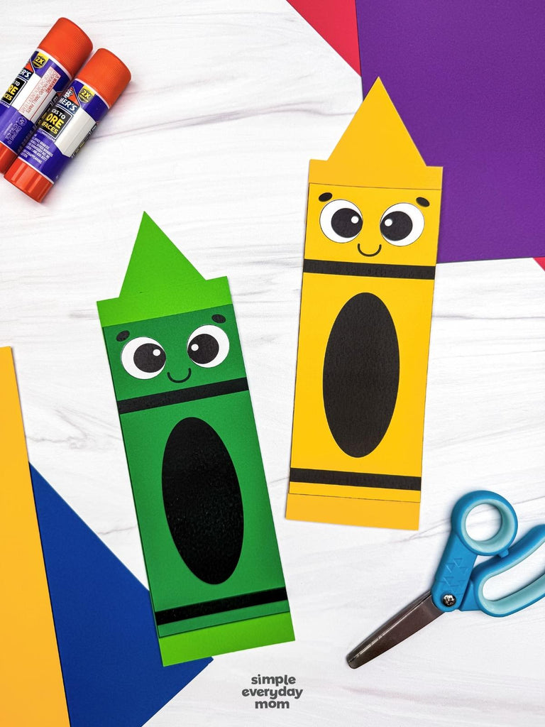 Two crayon-shaped paper crafts, one green and one yellow, with cartoon faces, displayed on a white wood background with craft supplies including glue sticks, scissors, and colored paper.