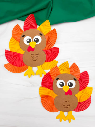 Two paper turkey crafts on a white background with green fabric.