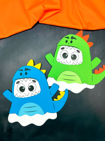 Two colorful paper dinosaur ghosts are disguised on a black surface with orange fabric in the background.