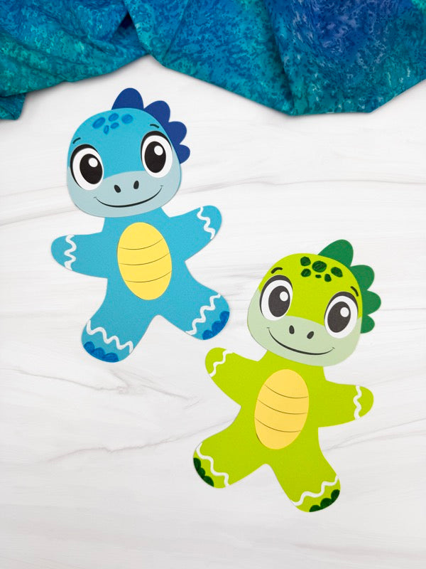 Two gingerbread men disguised as dinosaurs, one blue and one green, on a white background with a blue textured fabric in the background.
