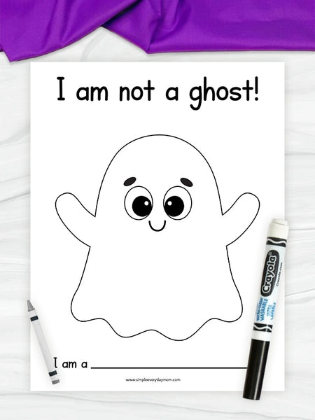 A printable coloring page featuring a cute black-and-white outline ghost with the text 'I am not a ghost!' and 'I am a ______'. The page is styled on a white wood surface with a purple fabric strip at the top, a crayon on the left, and a Crayola marker on the right.