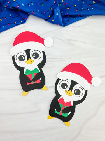 Two Christmas themed penguin crafts on white surface with blue fabric on top. 