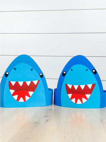 Two blue paper shark headbands with wide open red mouths showing white triangular teeth, cartoon eyes, and simple fin details, displayed on a light wood surface against a white shiplap wall.
