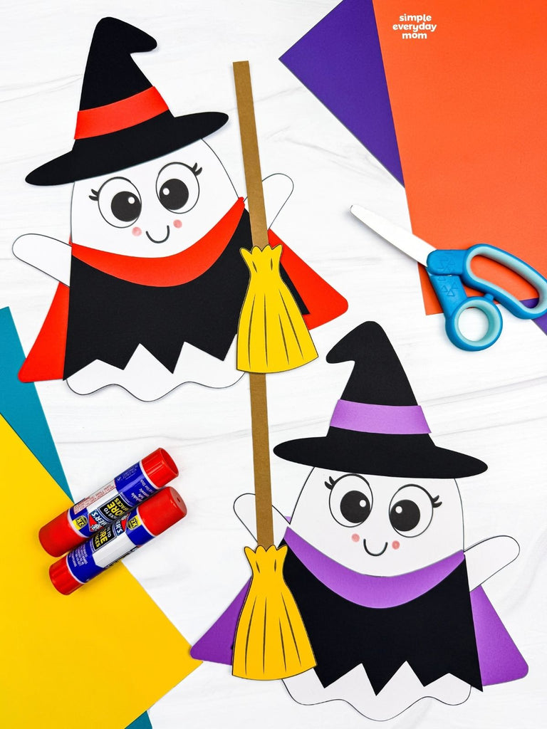 Two finished witch ghost disguise paper crafts on a white tabletop, featuring black pointed hats, matching capes, and yellow brooms. One ghost has red costume accents, while the other has purple accents, with colorful construction paper, scissors, and glue sticks nearby.