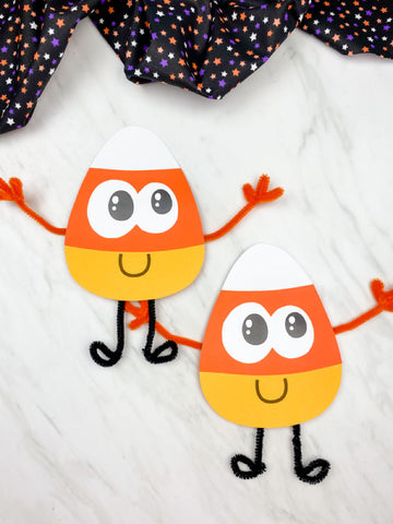 Two paper candy corn characters on a white background with a Halloween-themed fabric in the background.