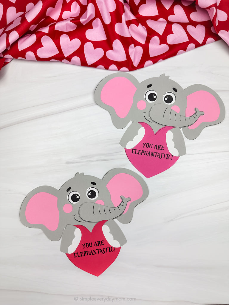 Two elephant-shaped crafts with pink hearts and text on a white surface with a heart-patterned fabric in the background.