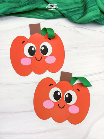 Two cute orange paper pumpkin decorations with smiling faces, pink cheeks, and green stems, displayed on a white wood background with a green fabric banner at the top.