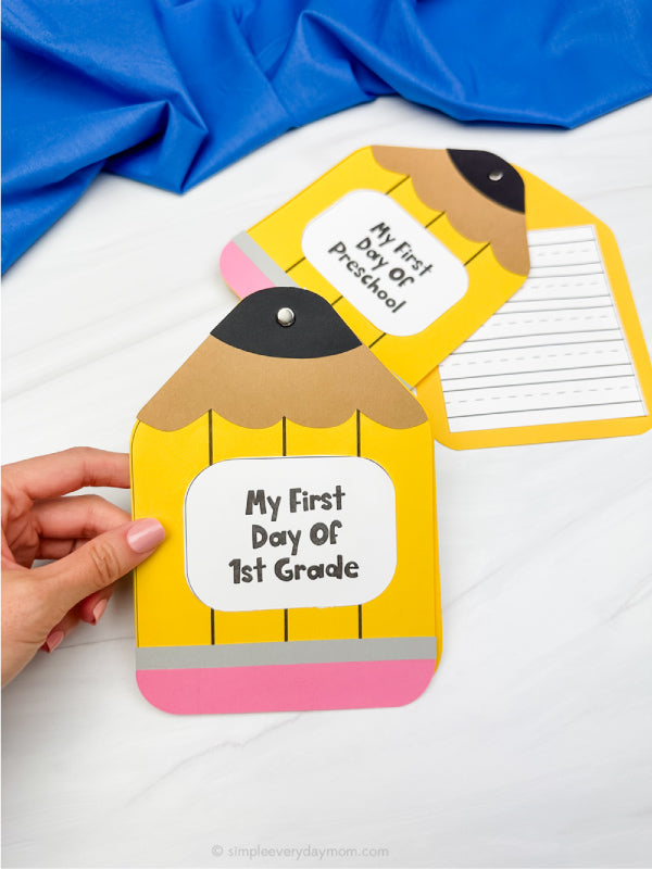 Hand holding a yellow school pencil-shaped card with 'My First Day of 1st Grade' text on a white background.