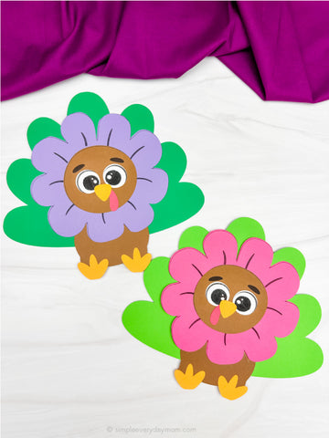 Two colorful paper turkey crafts disguised as flowers on a white background with a purple fabric.